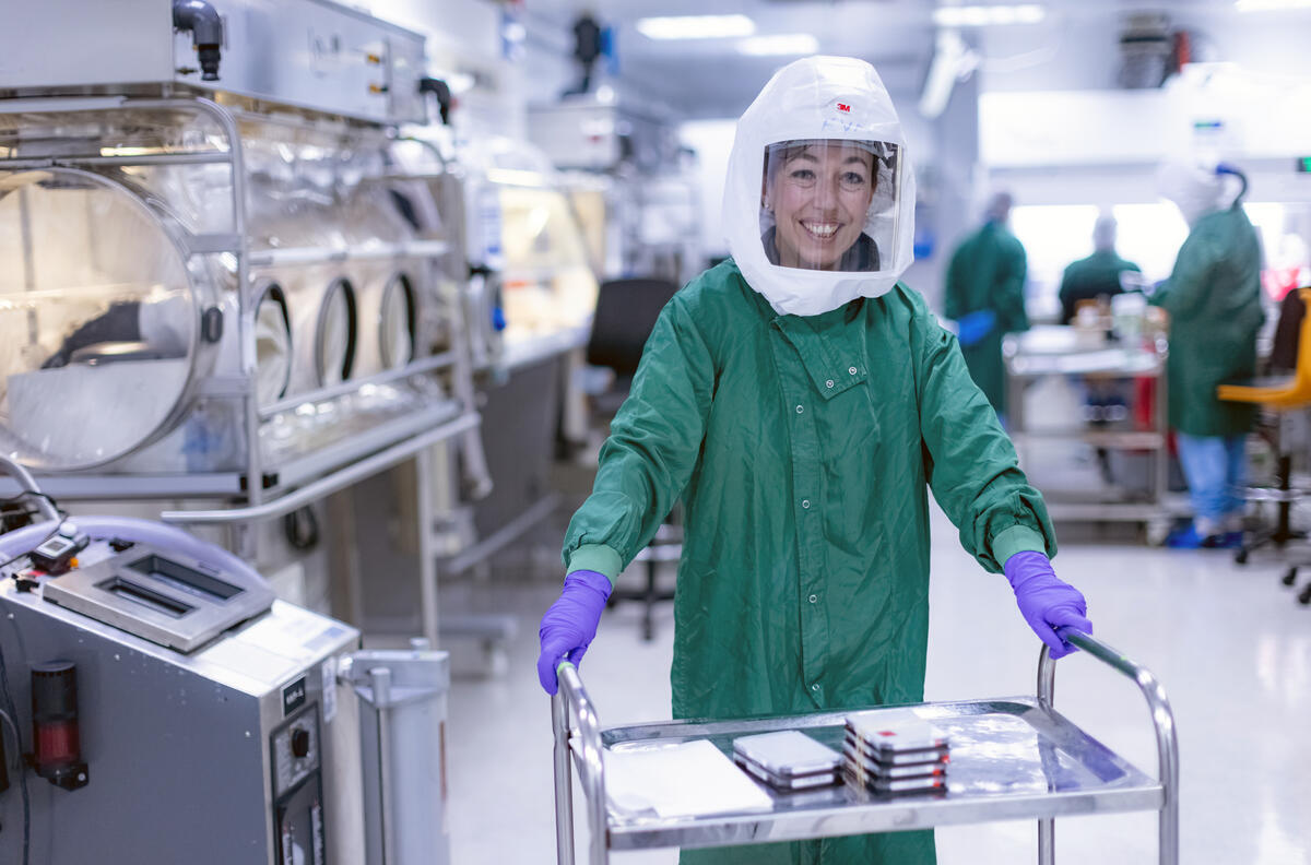 A scientist in a green gown, protective head visor and purple gloves pushing a trolley of lab equipment and smiling