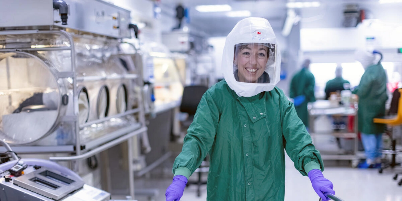 A scientist in a green gown, protective head visor and purple gloves pushing a trolley of lab equipment and smiling