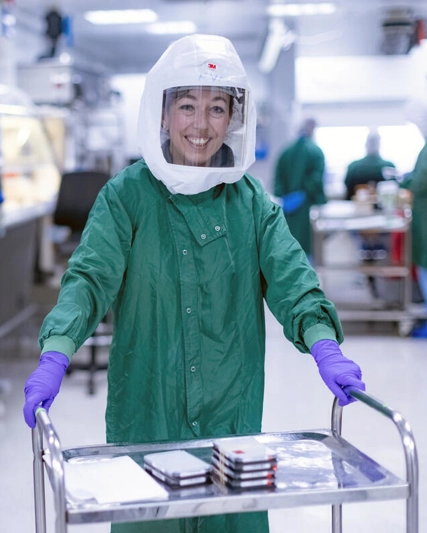 A scientist in a green gown, protective head visor and purple gloves pushing a trolley of lab equipment and smiling