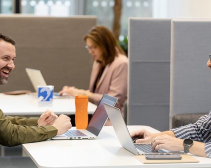 Two men using laptops laughing together, woman using laptop behind