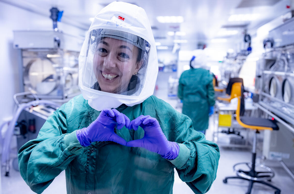 A scientist smiling and making a heart shape with their hands in purple gloves, a green gown and protective head gear
