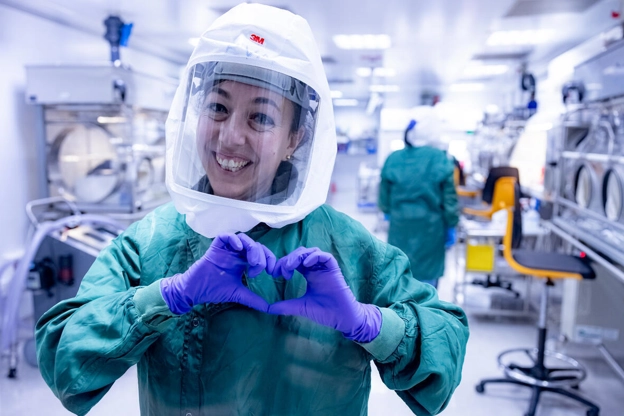 A scientist smiling and making a heart shape with their hands in purple gloves, a green gown and protective head gear