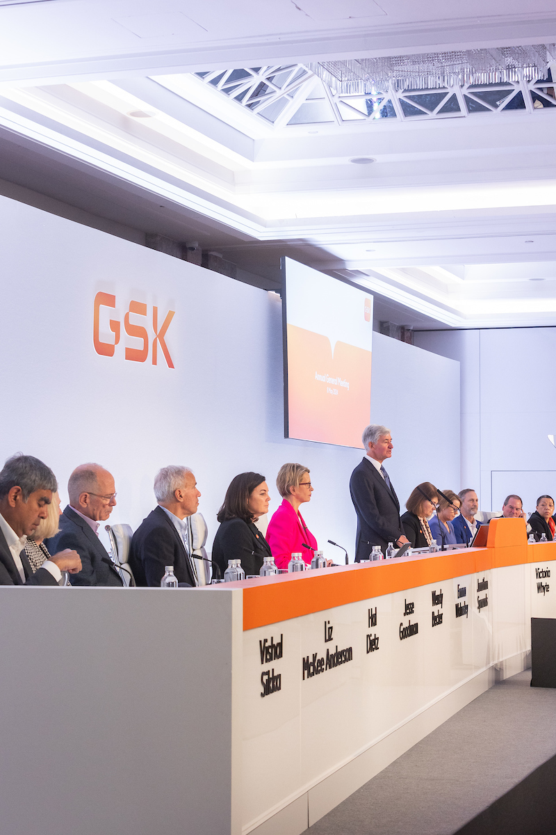 Board of directors, men and women, sitting on stage, listen to Chair Jon Symonds speaking at AGM