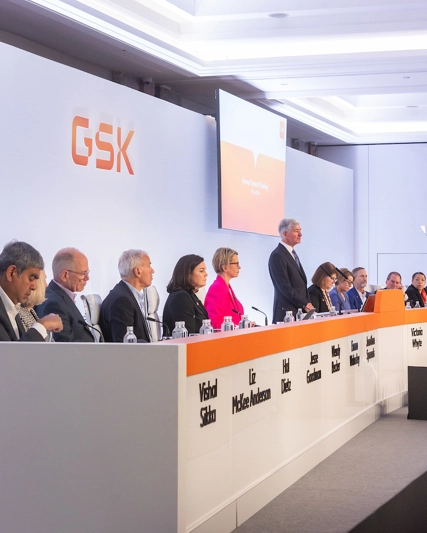 Board of directors, men and women, sitting on stage, listen to Chair Jon Symonds speaking at AGM